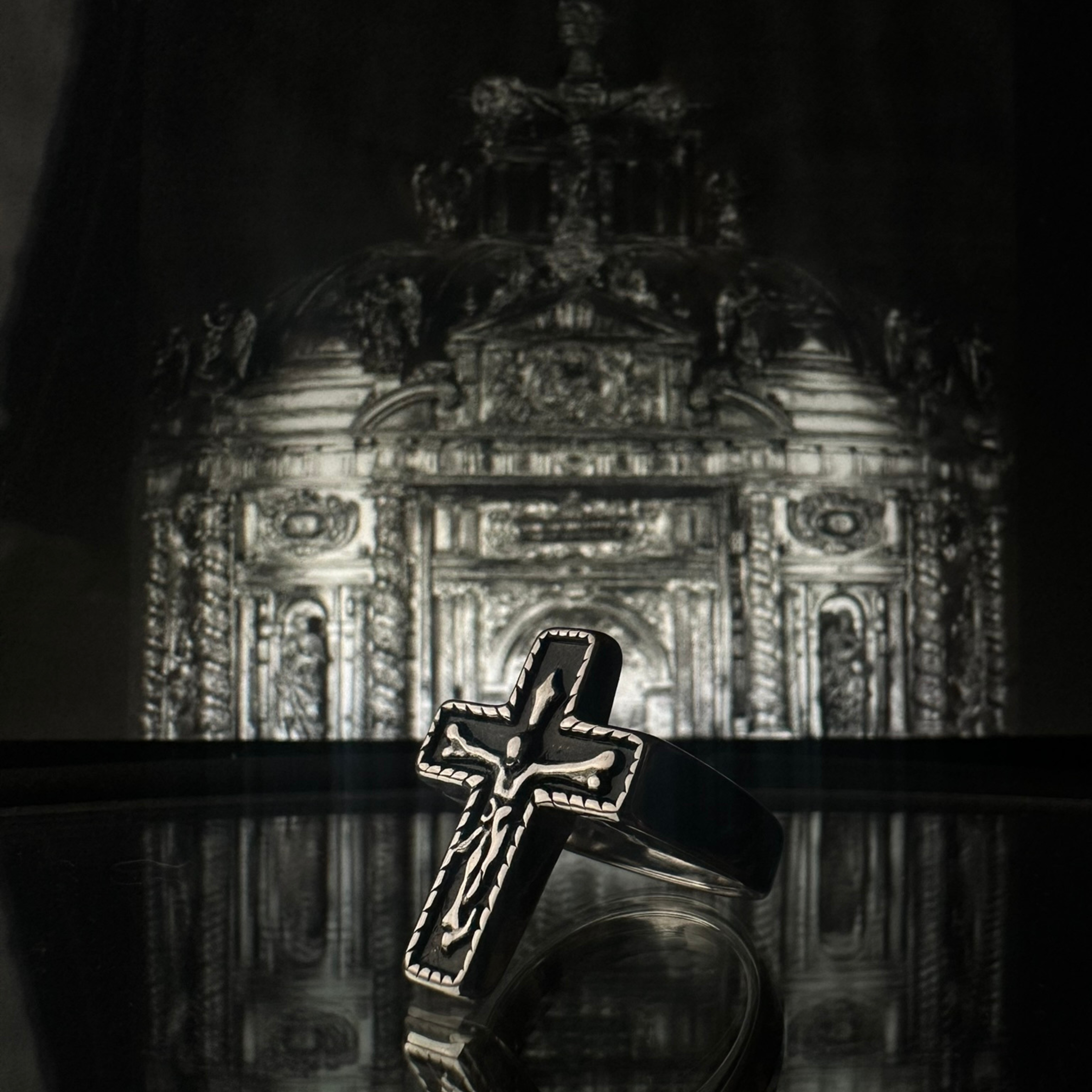 Anillo de acero inoxidable tipo sello 'Follow Ring Big Cross' con diseño de cruz en relieve, de la marca Follow God, presentado frente a un fondo arquitectónico clásico