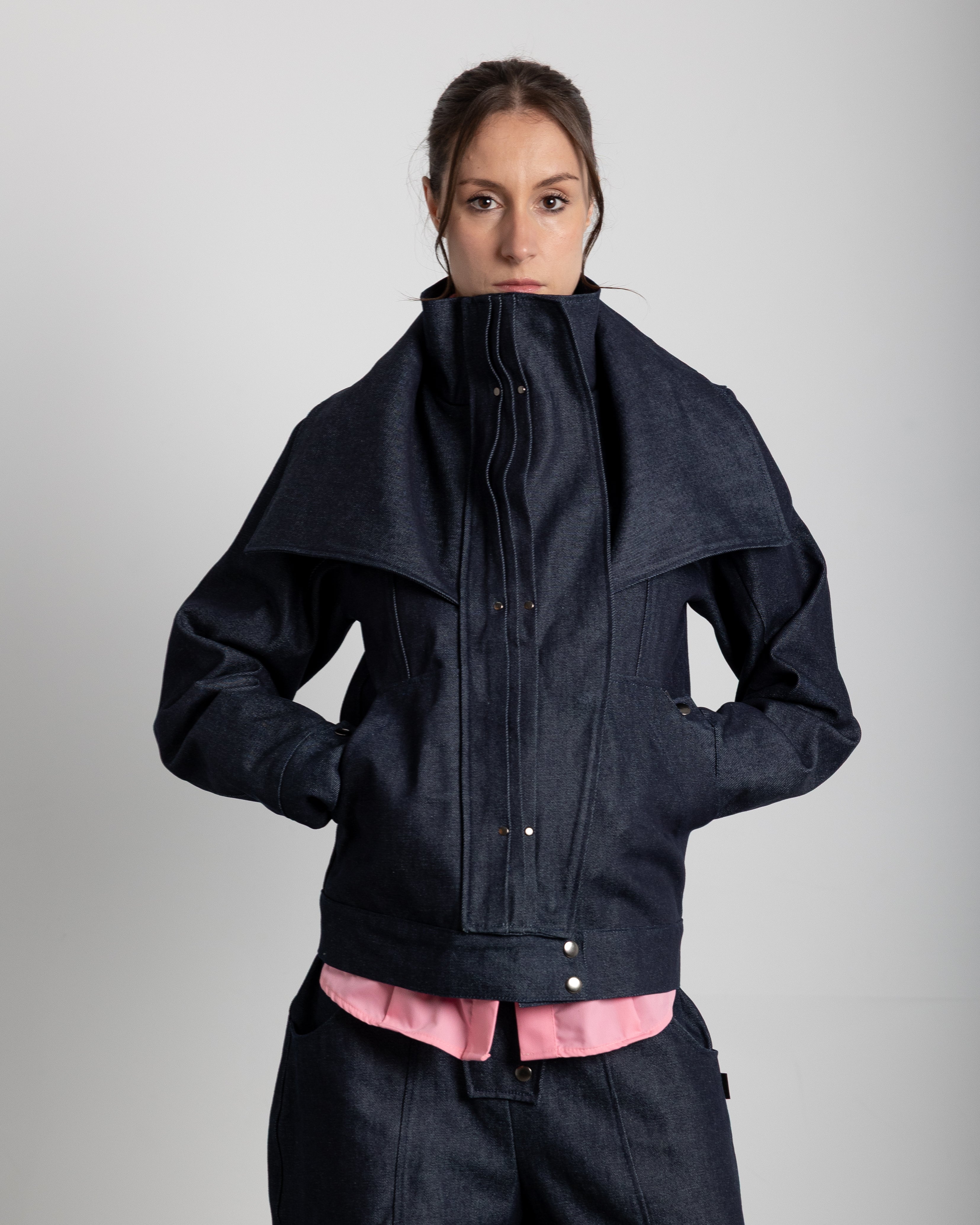 Chaqueta de capas en denim RAW 100% algodón. Chaqueta con cuello alto y solapas ajustables. Tapeta plisada con remaches plateados que cubren la cremallera. Bolsillos delanteros.